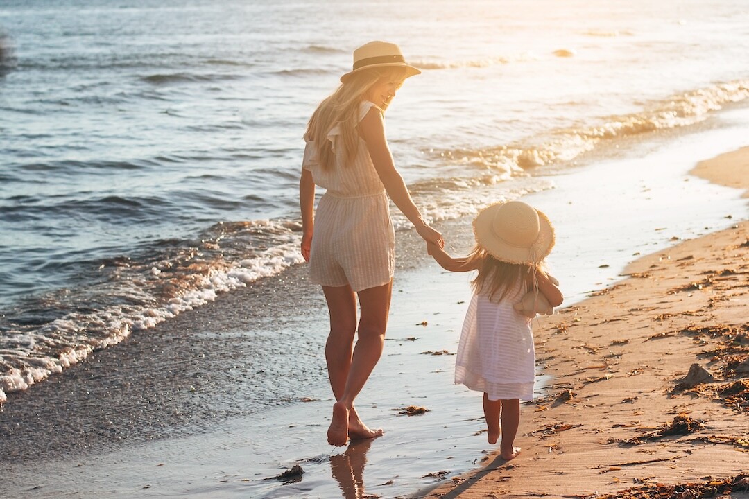 A woman and a child, both wearing summer outfits and wide-brimmed hats, walk hand-in-hand along a sandy beach at sunset. The waves gently lap at the shore, and the sky glows with warm, golden hues. The scene exudes tranquility and warmth.