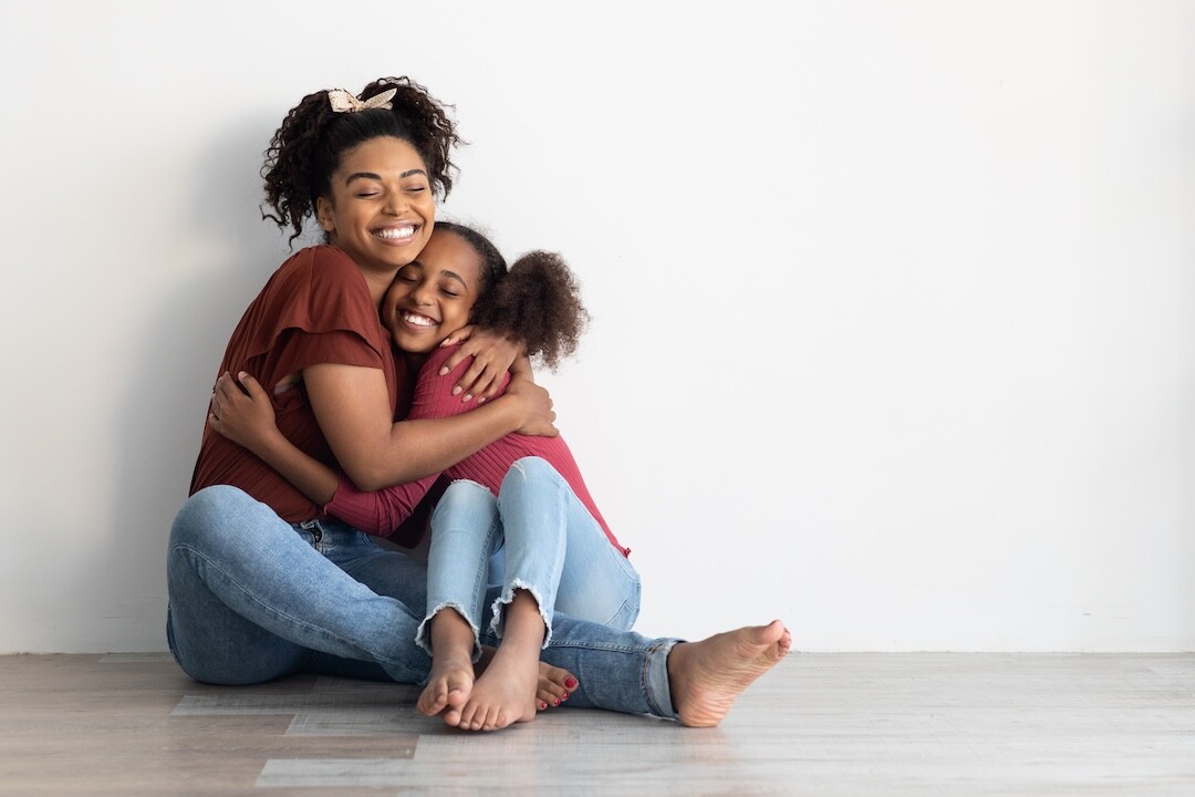 Two people sit on the floor in front of a white wall, smiling and hugging each other warmly. Both are dressed casually in jeans and t-shirts, appearing joyful and content. Their embrace showcases a close, affectionate relationship.