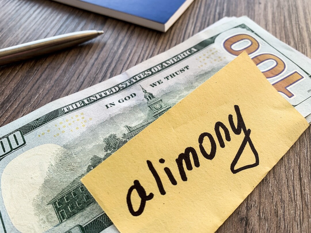A close-up image of a stack of 100-dollar bills with a yellow sticky note on top that reads "alimony." A pen and a notebook lie blurred in the background on a wooden surface.