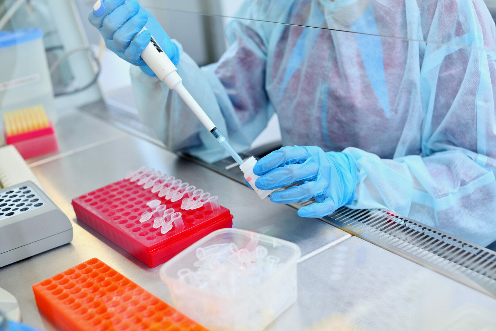 A scientist wearing protective gear and blue gloves uses a pipette to transfer a liquid sample into a small tube in a laboratory. The workspace is organized with red racks holding multiple small tubes and a clear container with additional tubes.