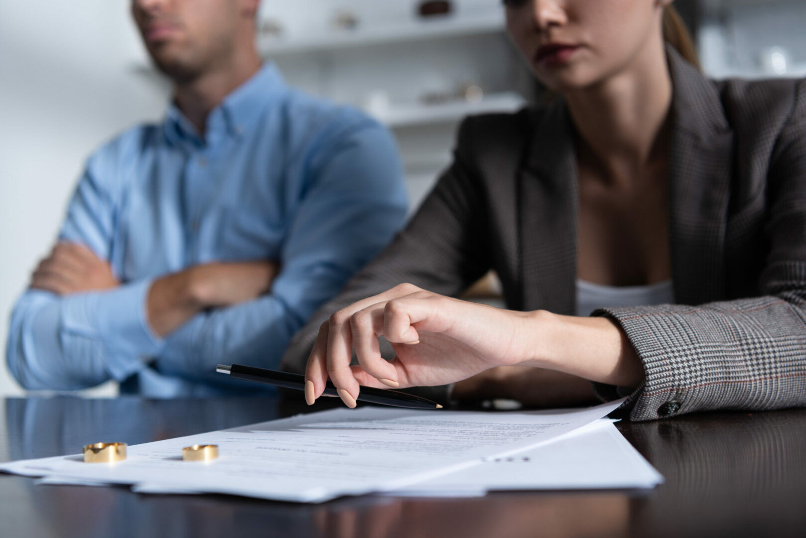 Two people, one in a blue shirt and the other in a grey blazer, sit at a table with documents and two wedding rings in focus. One person holds a pen, indicating they might be discussing or signing the papers. The mood appears serious and tense.