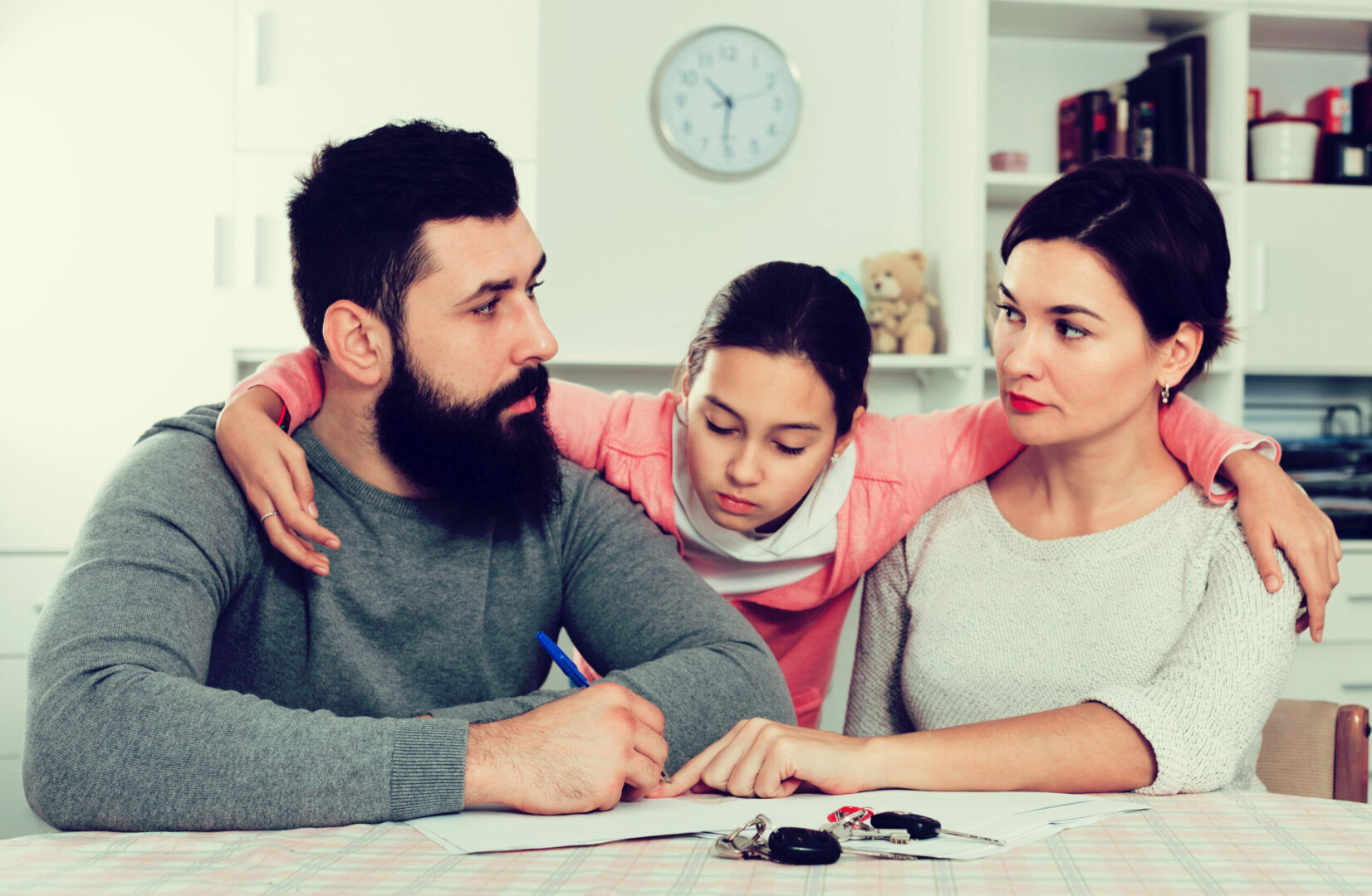 A family sits at a table with serious expressions. The father, with a beard, is writing on a piece of paper while the mother looks on. Their daughter, in a pink sweater, has her arms around both parents, leaning in to observe what her father is writing.