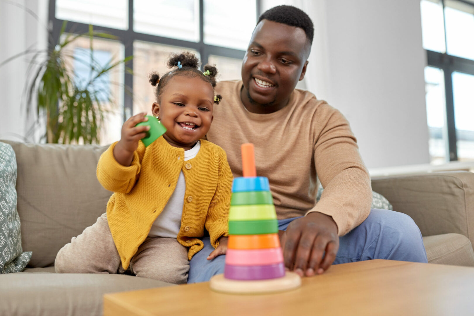 A father and his young daughter are playing with a colorful stacking toy while sitting on a couch. The girl, wearing a yellow sweater, smiles as she holds a green piece. The father, in a beige sweater, looks on lovingly. Large windows and indoor plants are in the background.