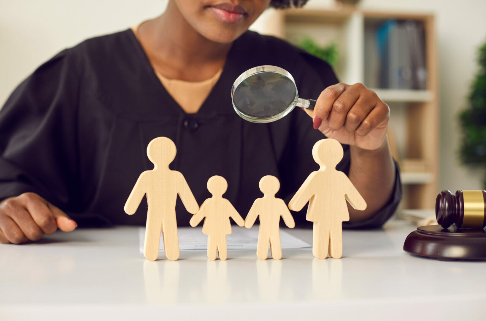 A person in a black robe holds a magnifying glass over wooden cutouts of a family of four on a desk. A gavel and books are in the background, suggesting a legal or judicial setting.