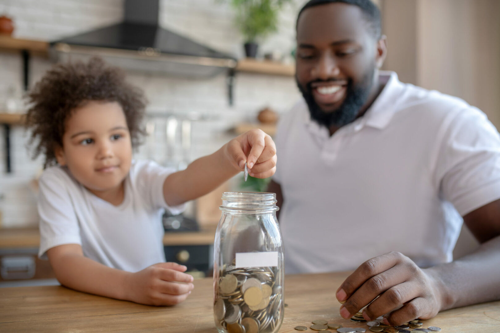 A man and a child, both wearing white shirts, are sitting at a table in a kitchen. The child is placing a coin into a clear glass jar filled with coins, while the man smiles and holds the jar steady. The background shows kitchen shelves and appliances.