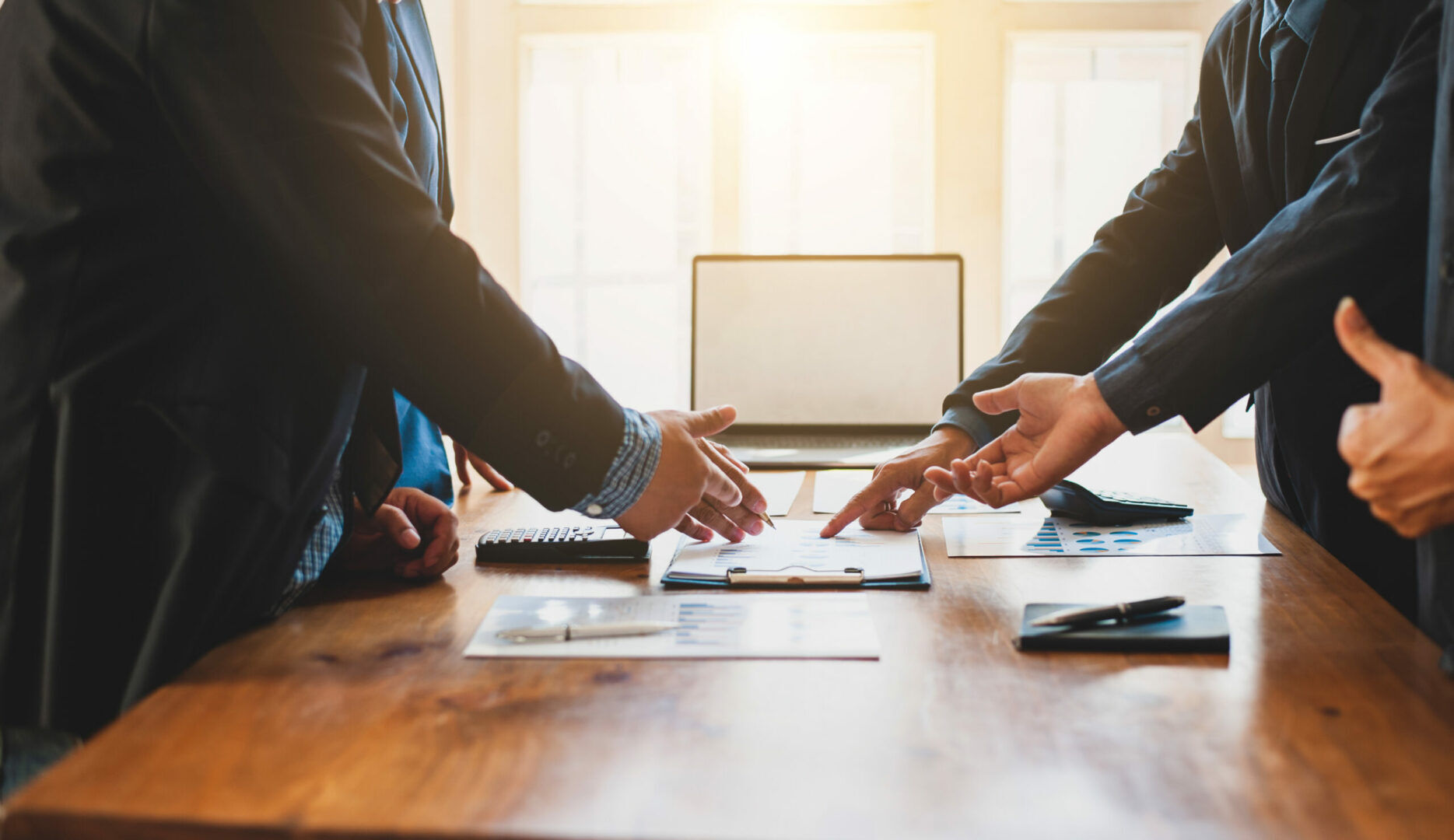 Business meeting with three people in suits discussing documents on a wooden table. A laptop, notebook, and charts are visible. One person extends a hand for a handshake, highlighting collaboration. Bright light streams through windows in the background.
