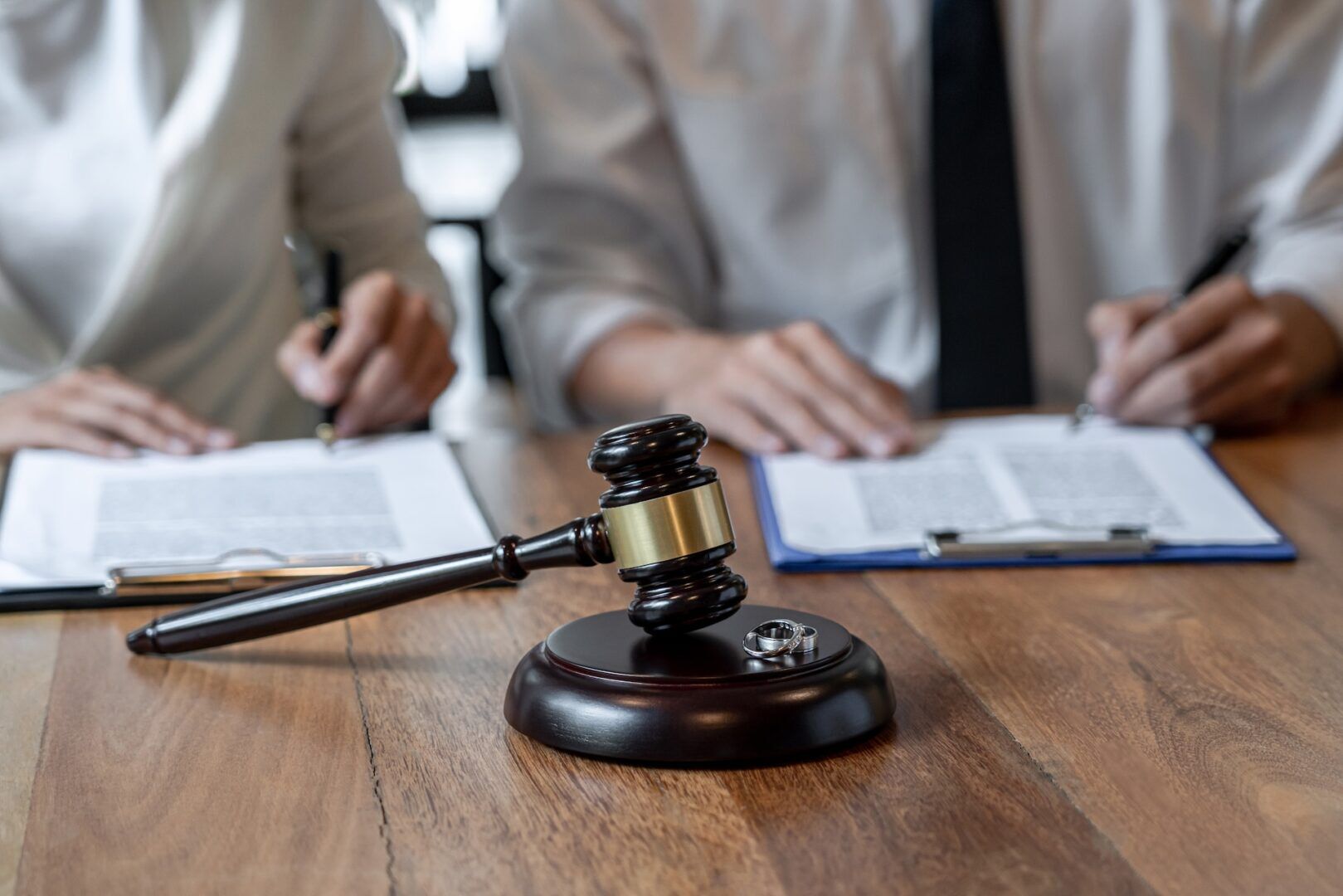 A gavel rests on a wooden table with two people sitting behind it. They are filling out paperwork on clipboards, suggesting a formal or legal setting. The individual on the left is dressed in white, and the figure on the right is wearing a shirt and tie.