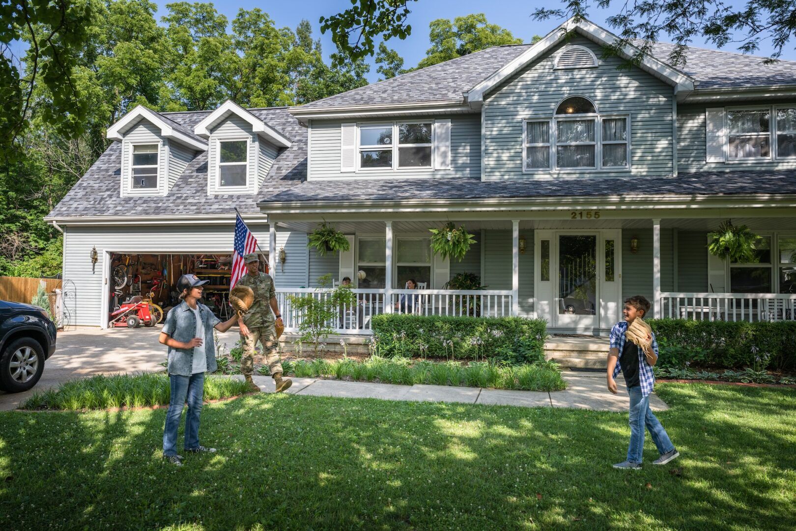 A soldier dressed in camouflage arrives at a blue-gray house with a large front lawn, while two children run towards him. An American flag is visible near the garage, and the porch is adorned with hanging plants and comfortable seating. Trees are in the background.