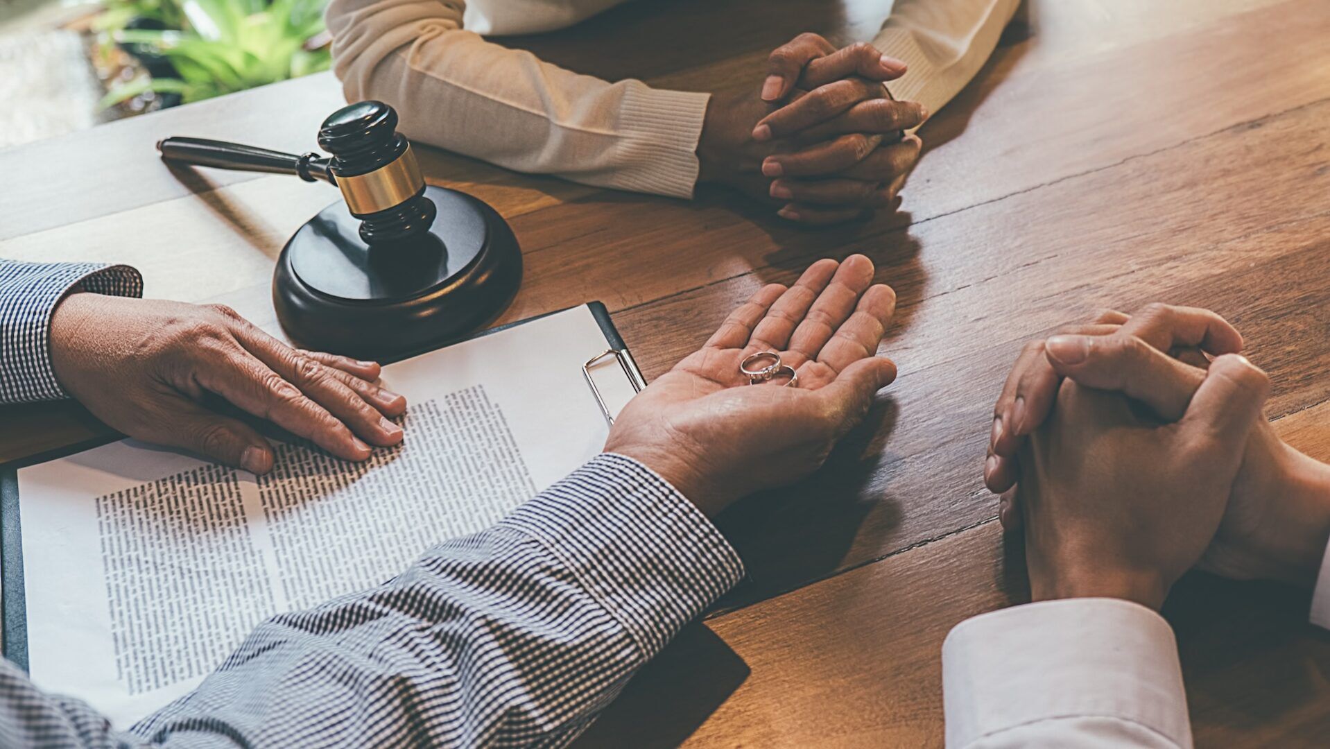 Two people sit across from each other at a wooden table, hands clasped. A legal document on a clipboard lies between them, next to a gavel. A third person extends a hand holding two rings, suggesting a discussion about divorce or separation.