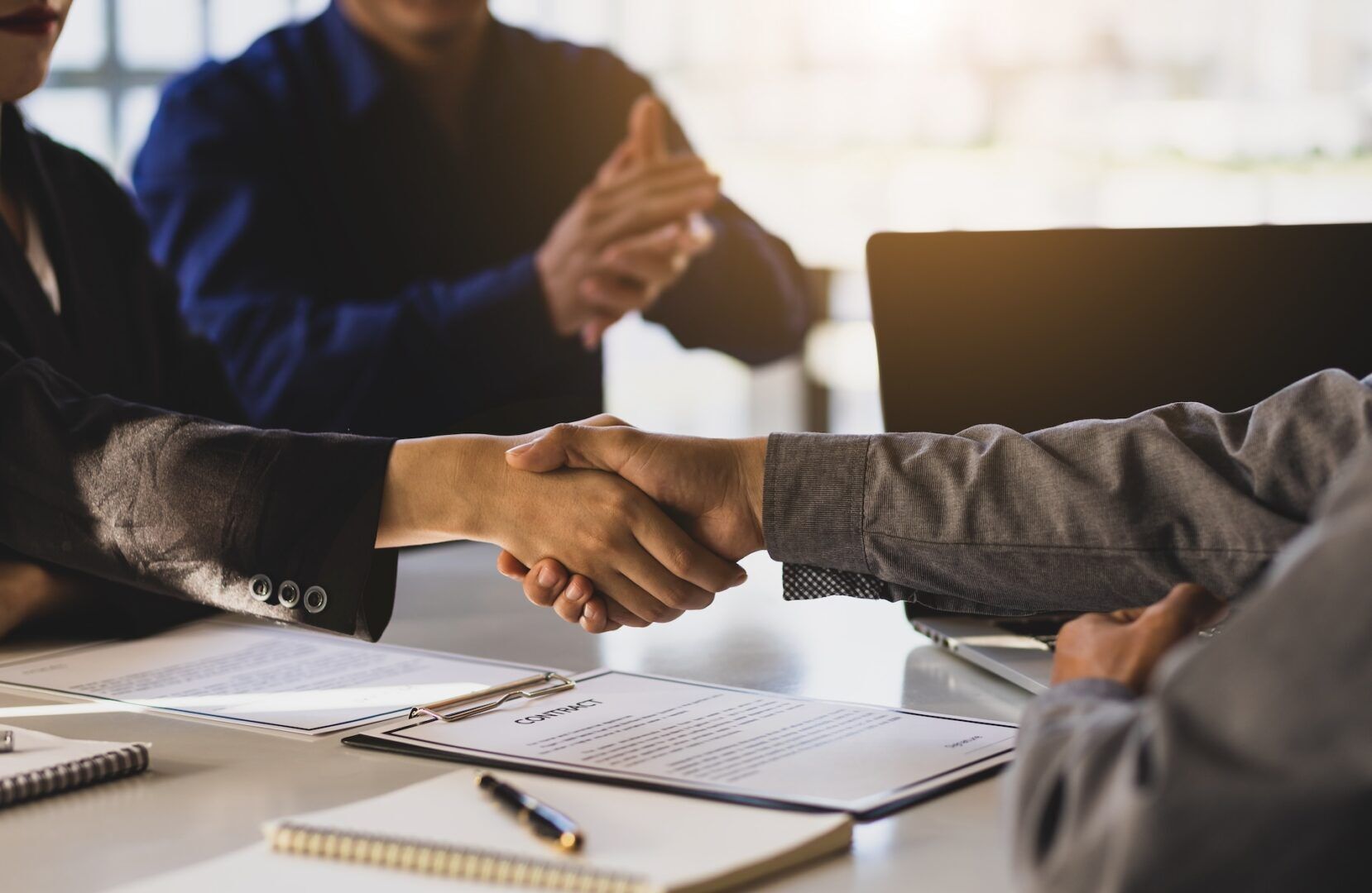 Two individuals are shaking hands across a table, presumably concluding a business deal. Documents and pens are on the table while a third person claps in the background. The setting appears to be a professional office environment with soft natural light.