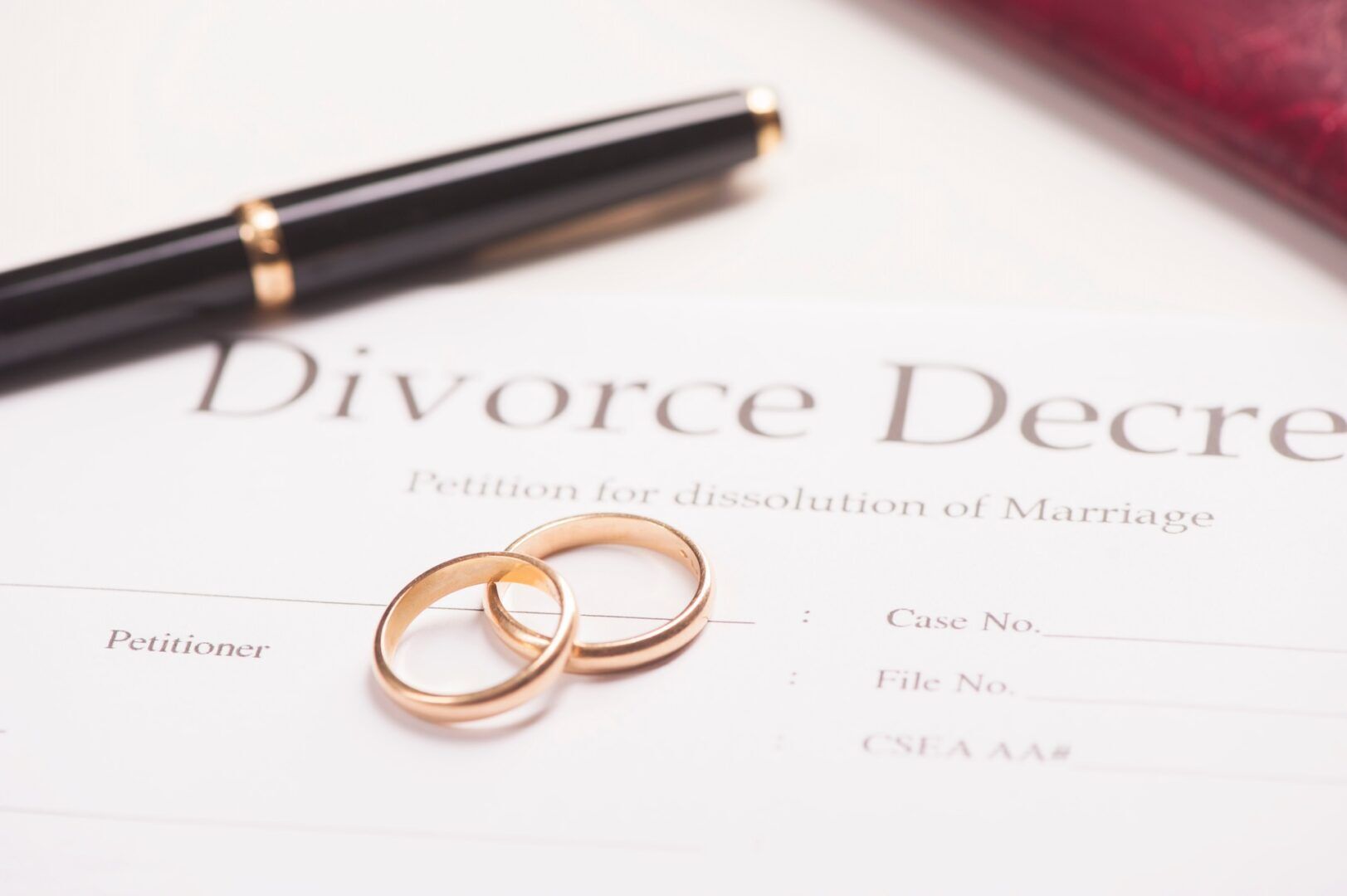Two gold wedding rings rest on a divorce decree document with a section labeled "Petition for dissolution of Marriage.” A black pen with gold detailing lies in the background, slightly out of focus.