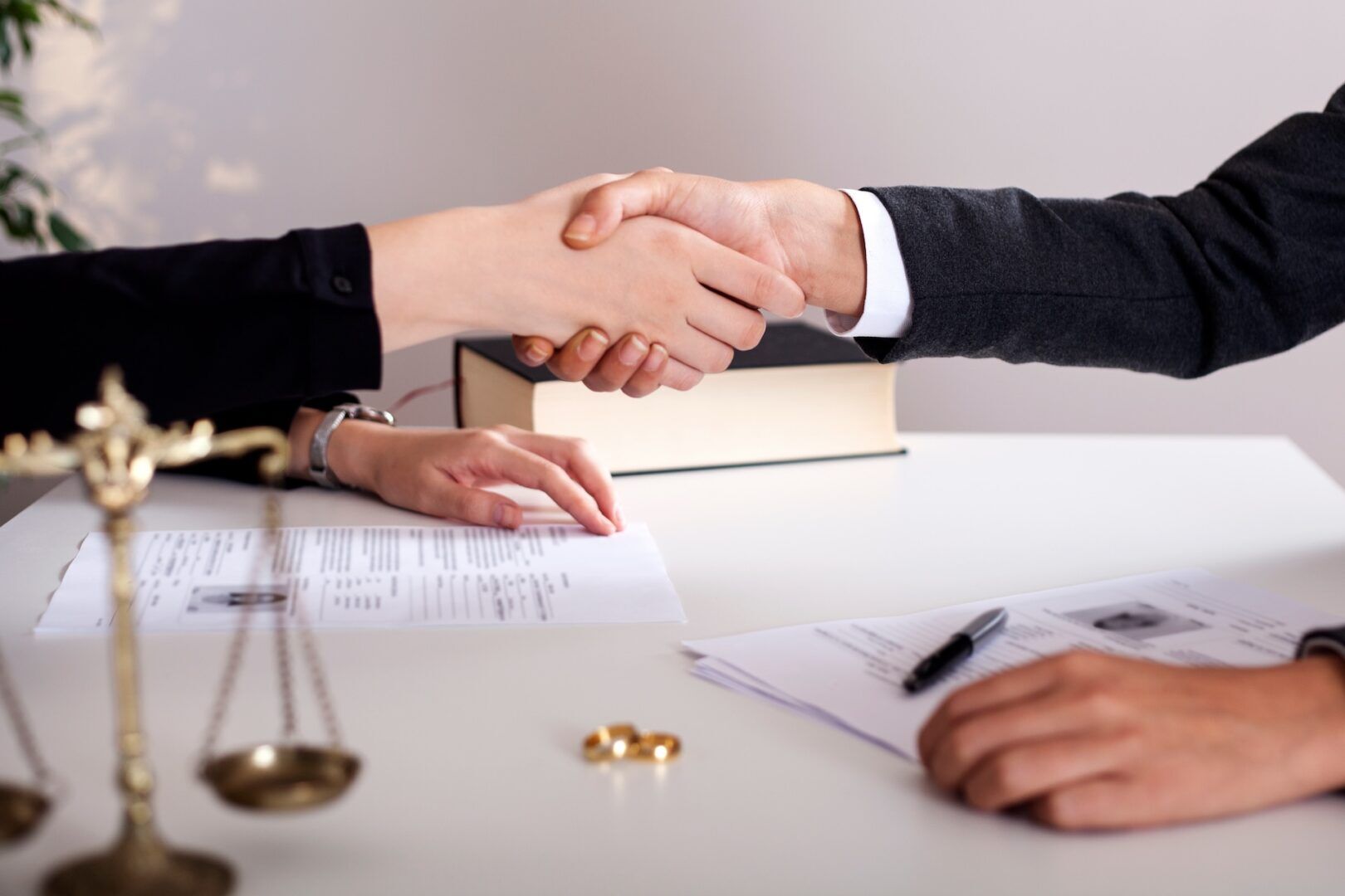 Two individuals are shaking hands across a white desk, signifying an agreement or conclusion of a deal. Documents are scattered on the desk along with a scale, symbolizing law or justice, and a pair of wedding rings.