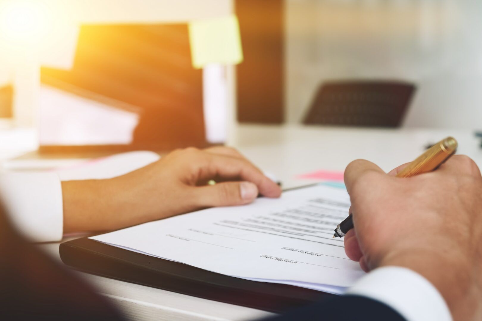 A close-up of hands signing a document on a clipboard. Blurred in the background is a laptop with sticky notes on the screen. Sunlight filters through the room, illuminating the workspace. The focus is on the pen and paper.