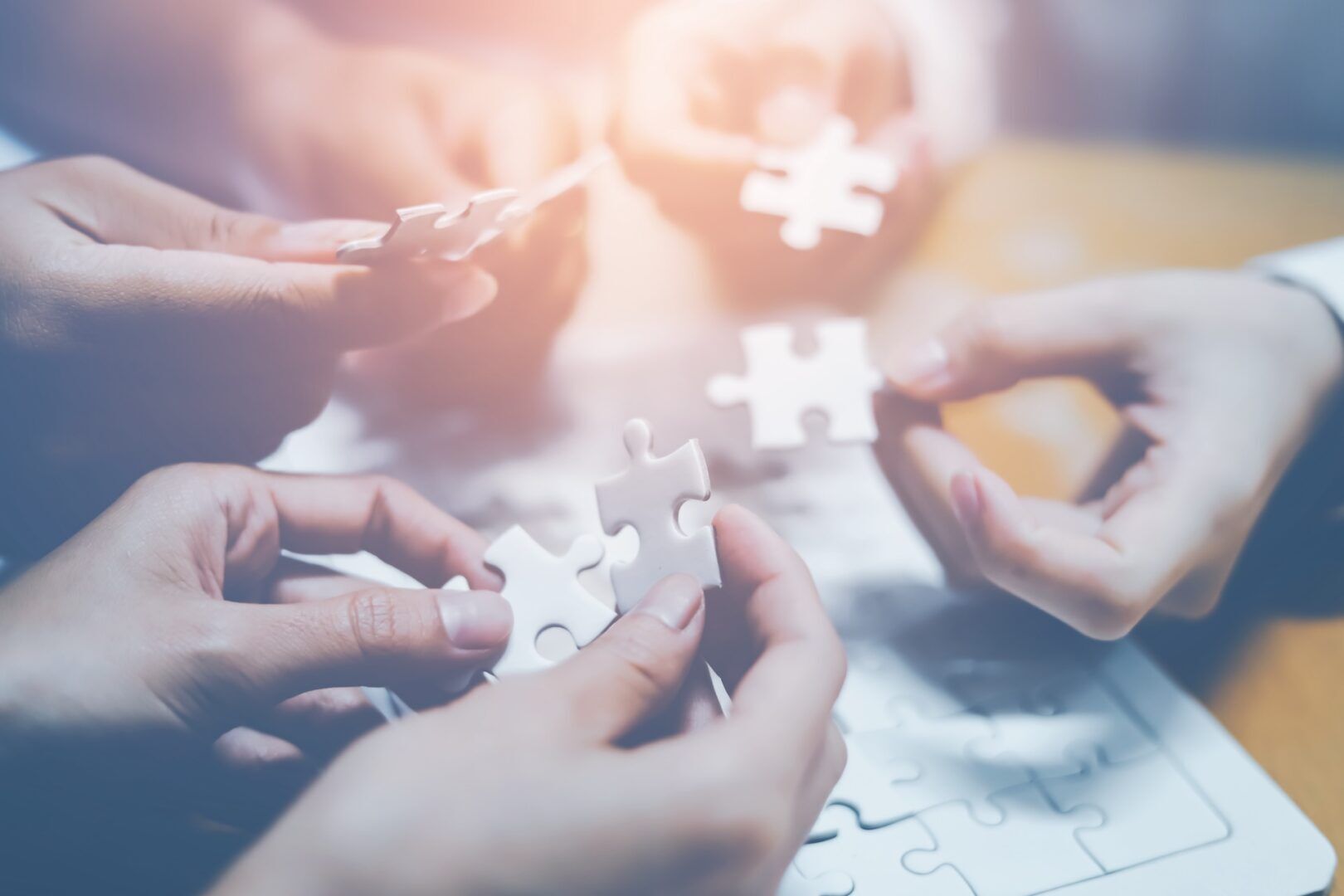 A group of hands collaborate to piece together a puzzle, with a bright light shining in the background. The image conveys teamwork, problem-solving, and collaboration.
