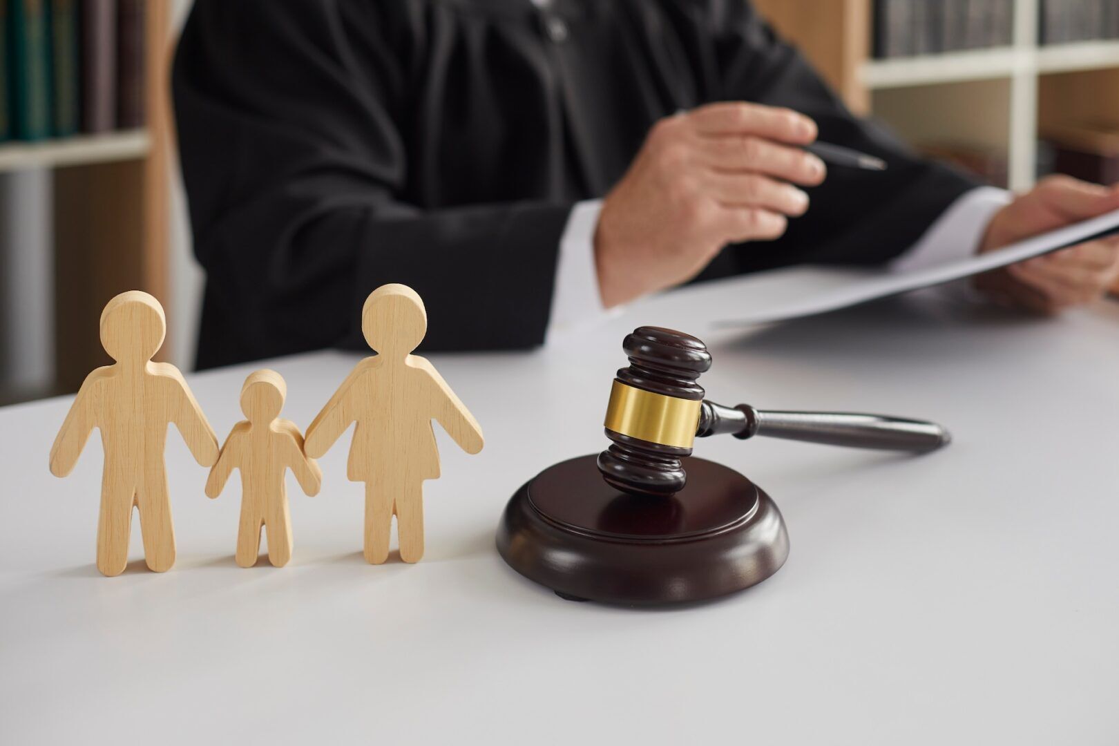 A judge in a black robe is holding a document, with wooden family figures and a gavel on a desk in the foreground. The family figures represent two adults and a child, symbolizing a family-related legal proceeding.