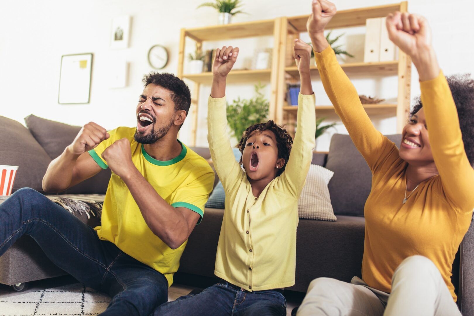 A family of three, dressed in yellow, sit on the floor in a living room, cheering enthusiastically. The father and child have their arms raised in excitement, while the mother also shows enthusiasm. Shelves with plants and books are in the background.