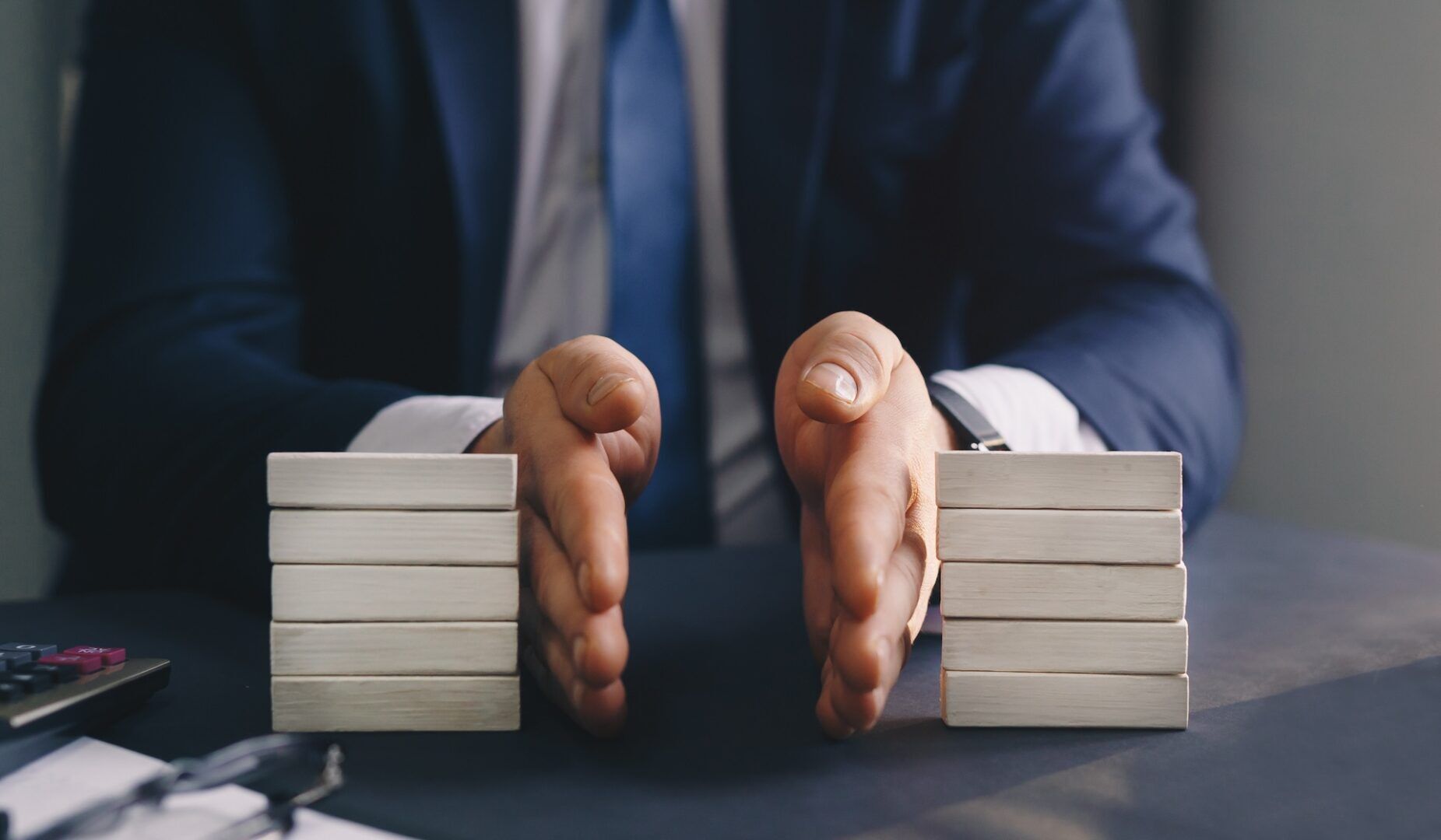 A person in a suit sits at a table with their hands placed between two stacks of wooden blocks, as if dividing or organizing them. A calculator and a pen are visible on the left side of the table. The background is blurred.