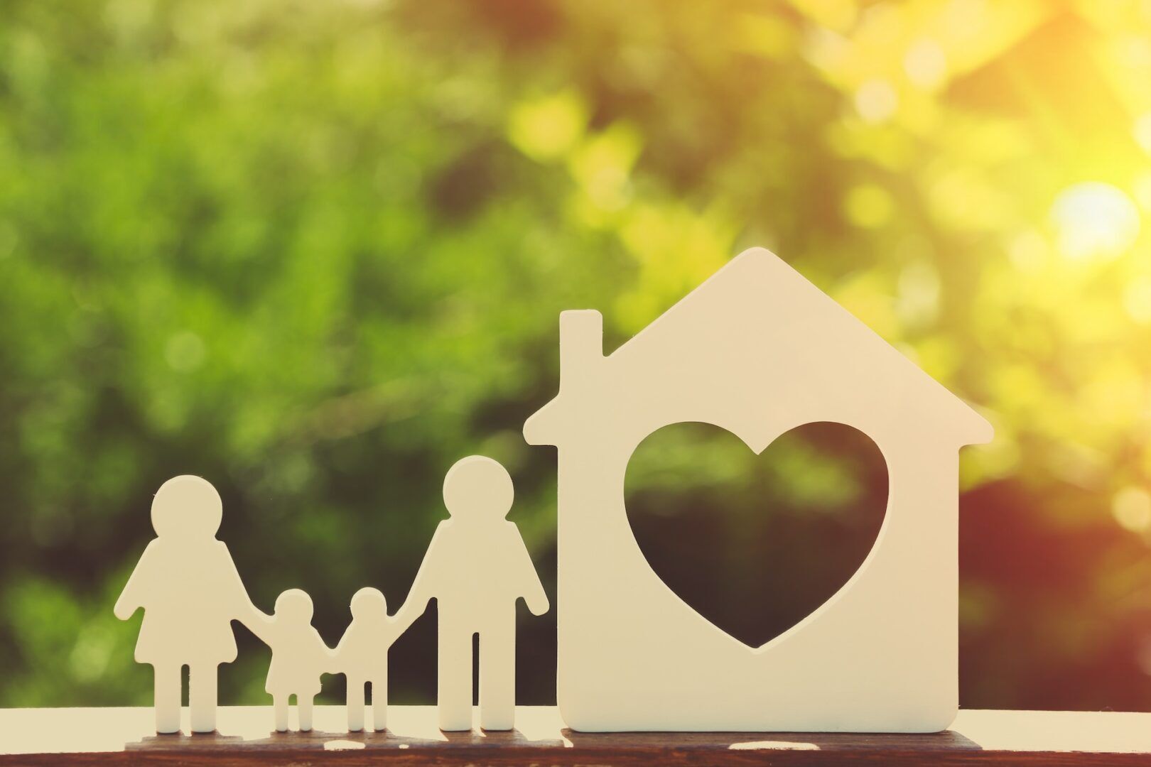 Silhouettes of a family holding hands, consisting of two adults and a child, stand next to a house shape with a heart cutout. The scene is set against a blurred, sunlit background of lush greenery.