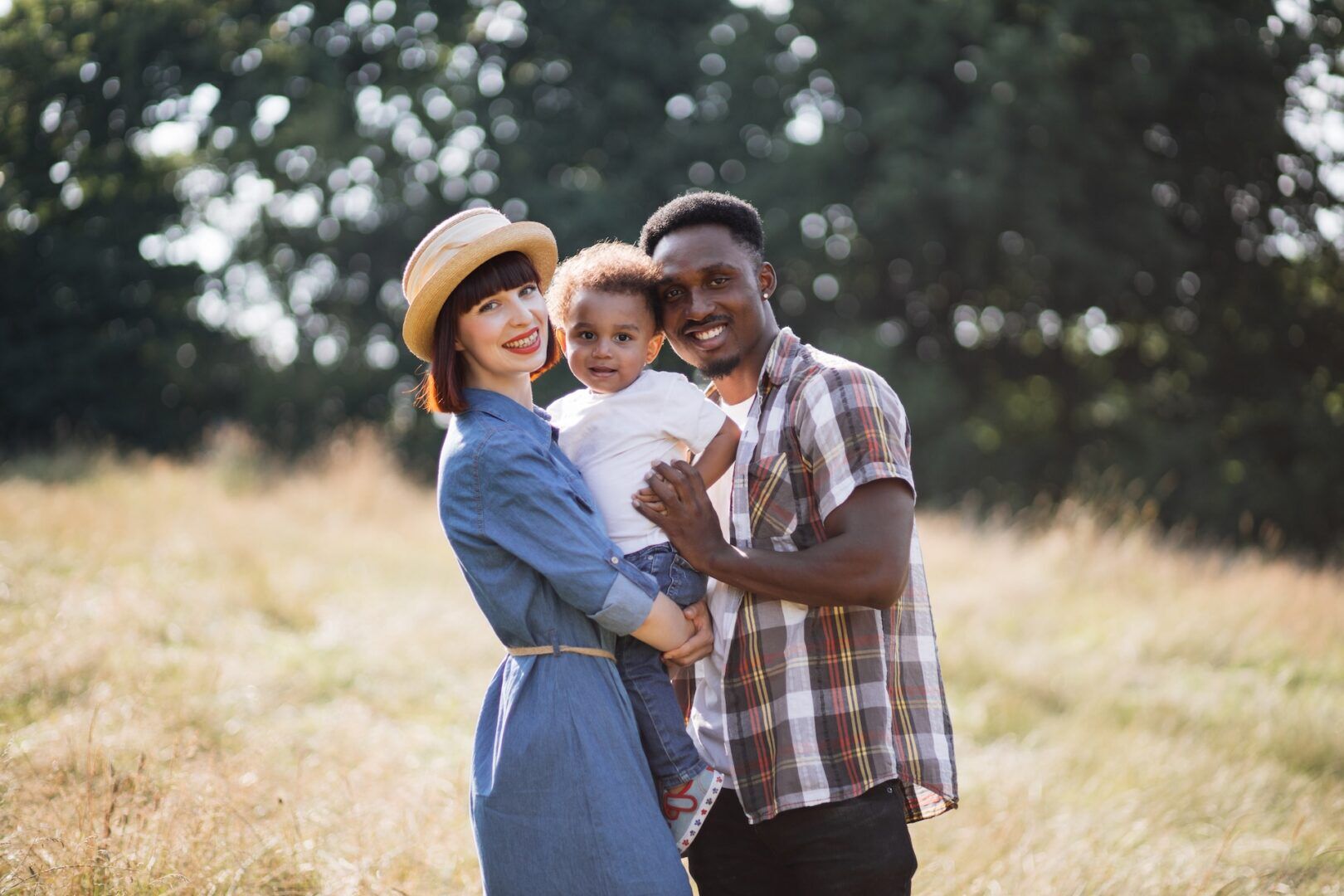 A happy family stands together in a sunny field. The mother wears a blue dress and straw hat, holding a toddler in a white shirt. The father is beside them in a plaid shirt. Trees are in the background, and they are all smiling.