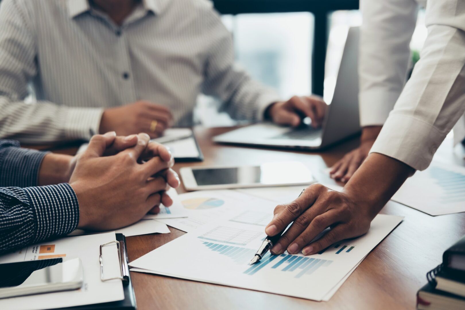 A group of people in business attire are seated around a table, reviewing documents and charts. One person is pointing at a graph on a page, while others have laptops and notepads in front of them. The scene suggests a meeting or discussion.
